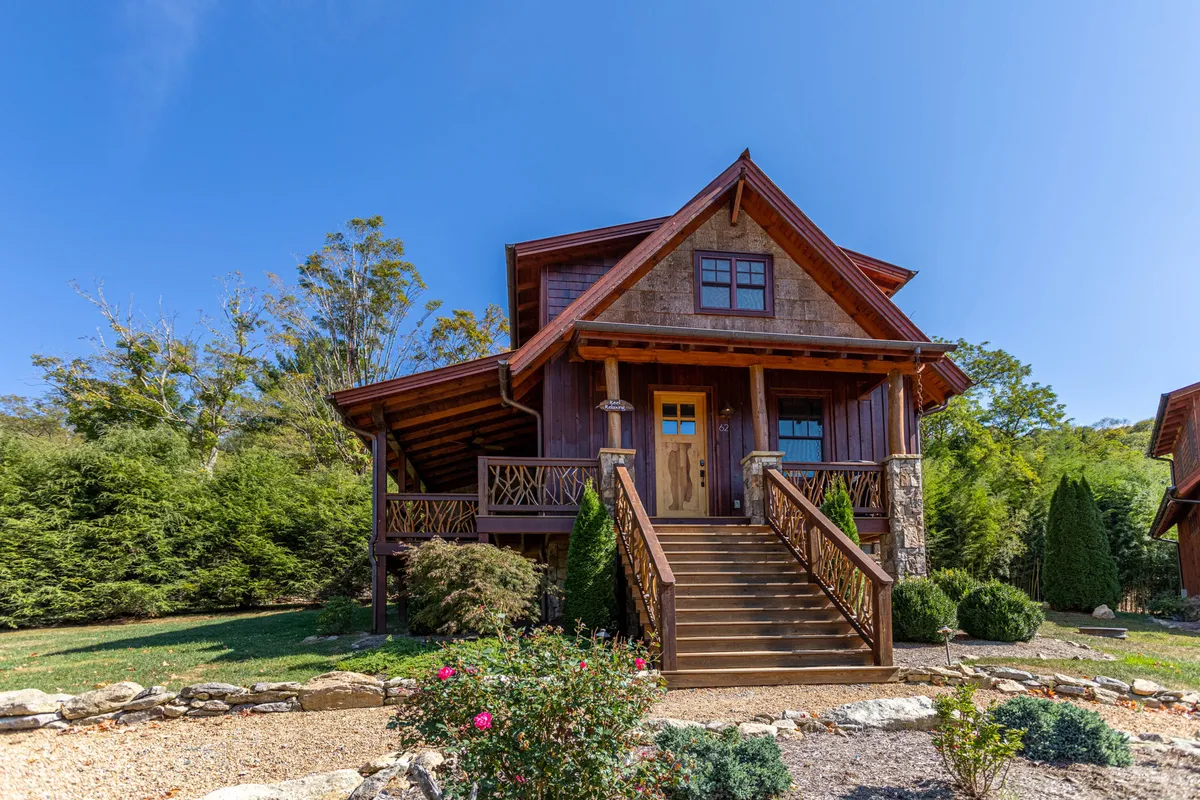 A welcoming wooden entrance door surrounded by greenery and flower beds, with steps leading up to the porch. The scene evokes a sense of arrival and connection between indoor comfort and outdoor adventure.