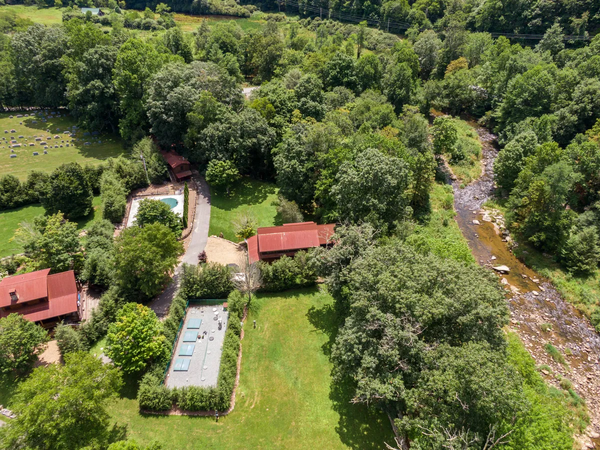 An aerial view of the property reveals lush greenery, a meandering stream, a swimming pool, and spaces where children are seen playing in the yard. The tranquil setting includes areas perfect for family picnics and relaxation by the water, blending luxury with the great outdoors.