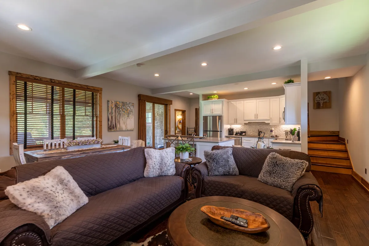 A view of the main living area that captures comfortable sofas, the dining table, kitchen in the background, decorative artwork, and a staircase. The space feels cozy and conducive to socializing, dining, and relaxing.