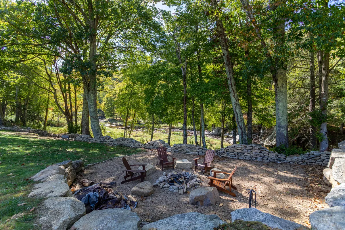 A cozy fire pit encircled by Adirondack chairs is set within a wooded area, surrounded by trees and a rustic stone wall. The inviting setup hints at evenings spent stargazing, sharing stories, or sipping wine by the fire.