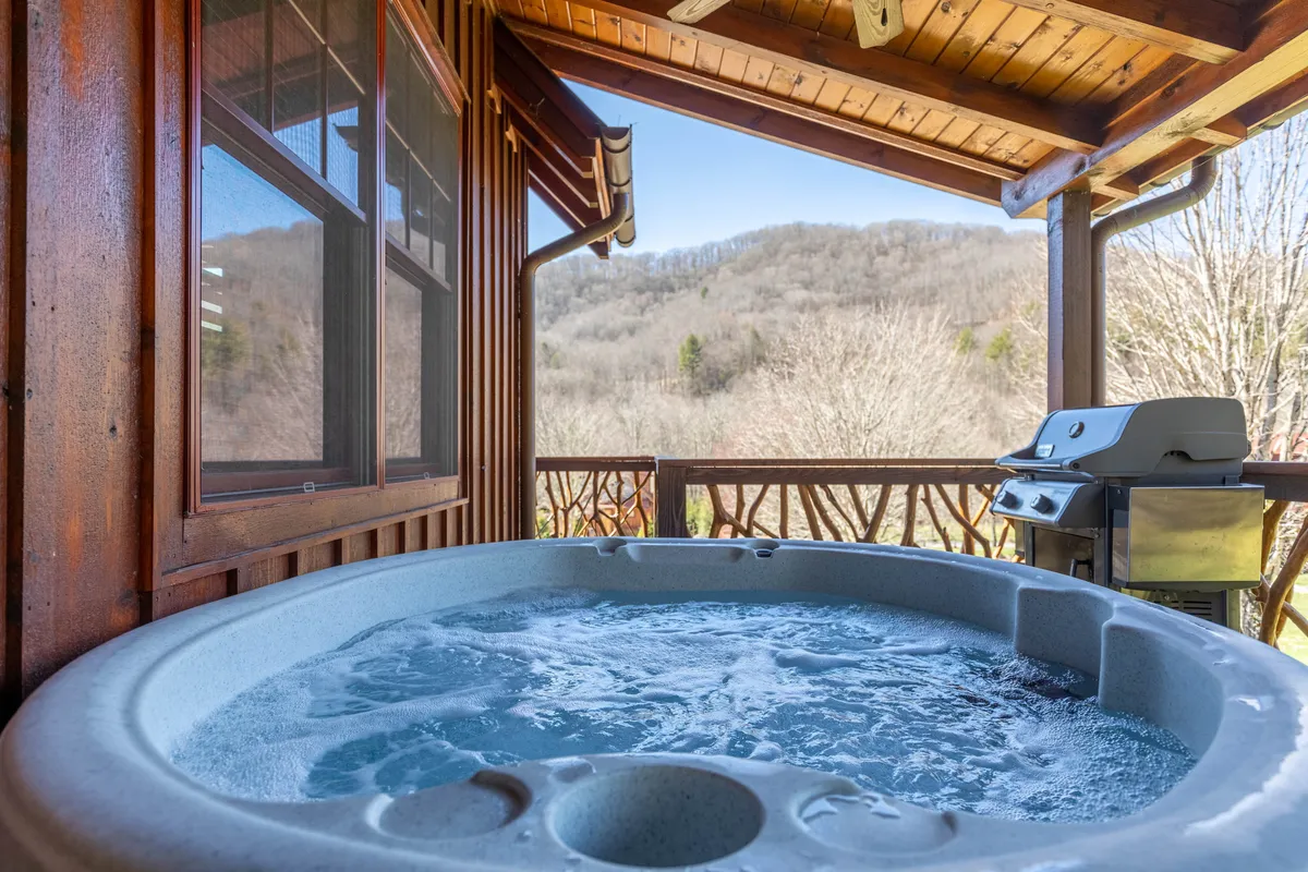 This exterior image features a private hot tub set on a deck with panoramic mountain views, capturing the tranquil ambiance of soaking amidst nature. A grill is also visible, suggesting added relaxation and leisure options outdoors.