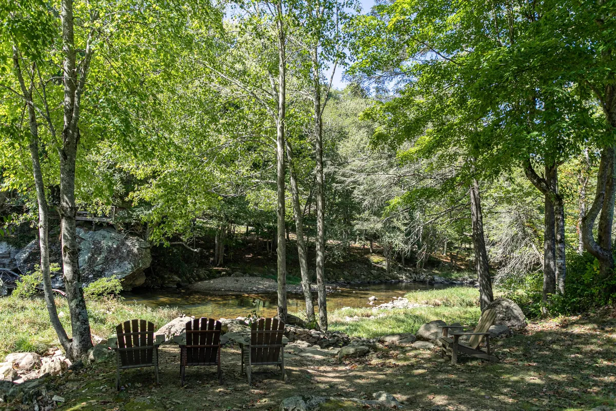 Three wooden chairs are arranged on the grassy bank of a stream, surrounded by lush greenery. The scene exudes intimacy and seclusion, suggesting a perfect spot for couples to relax with morning coffee or enjoy an evening by the water.