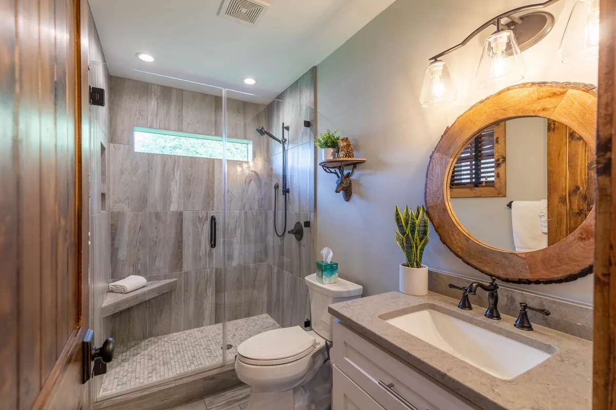A stylish, modern bathroom featuring a spacious shower area, a sleek round mirror above the sink, and a small shelf with a green plant, all presented in tranquil, natural tones.